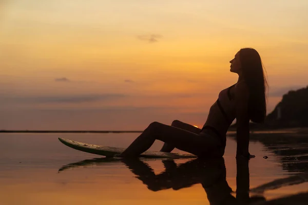 Silueta y reflejo de la niña sentada en la tabla de surf en la playa del océano en el fondo de la hermosa puesta de sol — Foto de Stock