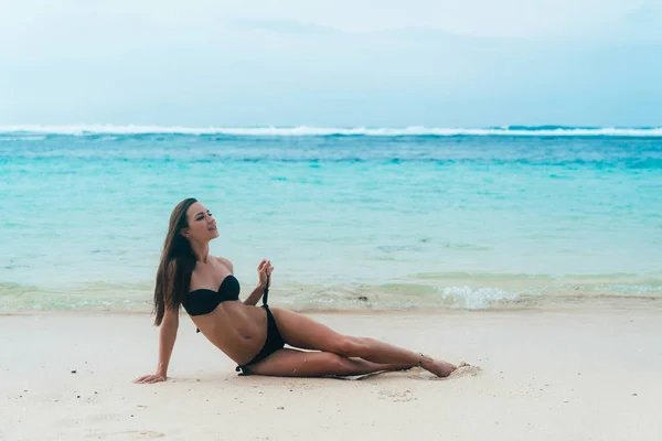 Sexy chica bronceada en traje de baño negro posando en la playa de arena cerca del océano. Hermosa modelo broncea y descansa — Foto de Stock