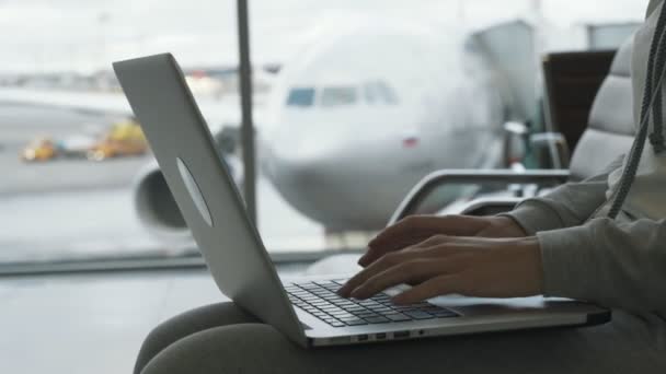 Руки крупным планом женщины, работающей на ноутбуке в зале аэропорта, на заднем плане самолета в окне ждут посадки — стоковое видео