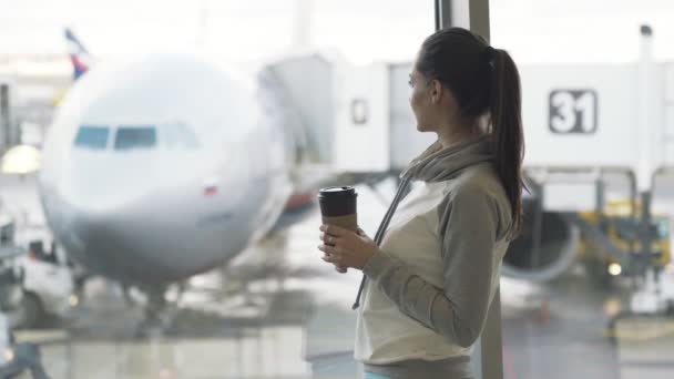 Gadis cantik dalam minuman sportwear kopi takeaway di bandara dekat jendela dengan pesawat di latar belakang — Stok Video