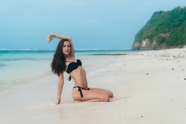 Sexy chica bronceada en traje de baño negro posando en la playa de arena cerca del océano. Hermosa modelo broncea y descansa — Foto de Stock