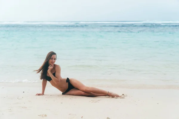 Szexi lány cserzett fekete fürdőruha pózol a homokos tengerpart közelében óceán. Gyönyörű modell sunbathes, és nyugszik — Stock Fotó