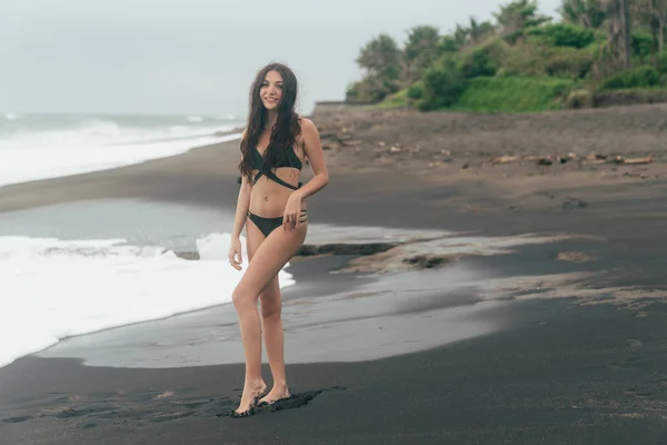 Bonne fille brune en maillot de bain posant sur la plage de sable noir. Modèle attrayant avec corps magnifique reposant près de l'océan — Photo