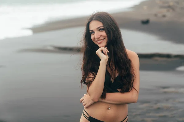 Güzel gülümseme ile mutlu kız portresi. Genç kadın siyah kum plajı üzerinde poz mayo — Stok fotoğraf