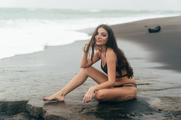 Attractive traveler in swimsuit sits on beach with black sand. Sexy girl spends vacation on ocean. — Stock Photo, Image