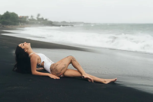 Sexy Mädchen mit perfektem Körper liegt am schwarzen Sandstrand. junge Frau genießt entspannende Zeit am Strand. — Stockfoto