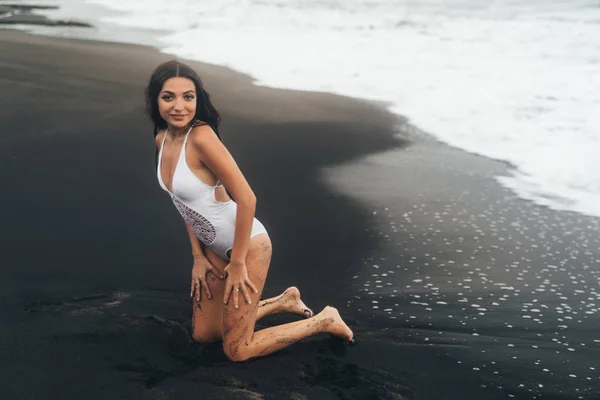 Ragazza carina in costume da bagno bianco in posa sulla spiaggia di sabbia nera. Modello sexy a riposo vicino all'oceano — Foto Stock