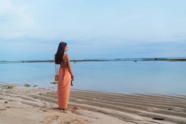 Arka görünümü ince kız sundress uzun kayış üzerinde çanta ile uzun saçlı kumlu tropikal kumsalda yürür