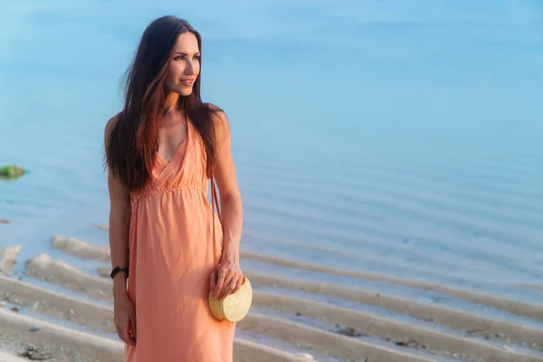 Muchacha atractiva con el pelo largo en vestido de sol con elegante bolsa de ratán de moda descansando en la playa — Foto de Stock