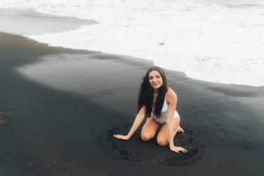 Okyanus dalgaları siyah kum plajda beyaz mayo siting içinde gülümseyen güzel kız