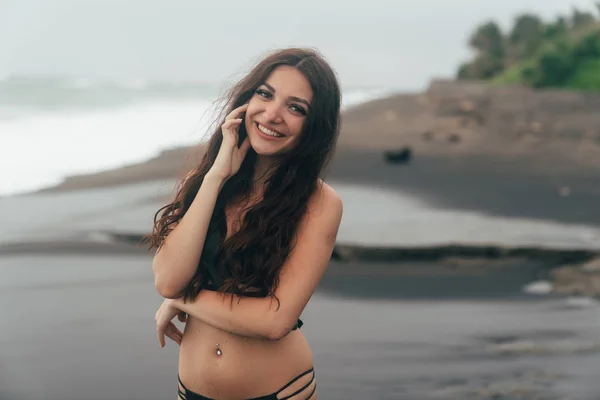 Porträt eines süßen Mädchens mit einem schönen Lächeln im schwarzen Badeanzug, das am Strand posiert. Reisende verbringen Zeit an der Küste. — Stockfoto