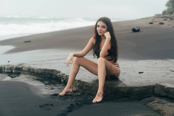 Schönes Mädchen mit langen Haaren im Badeanzug am schwarzen Sandstrand posierend — Stockfoto