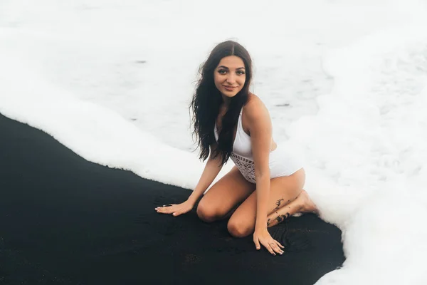 Beautiful smiling girl in white swimwear siting on black sand beach in waves of ocean — Stock Photo, Image