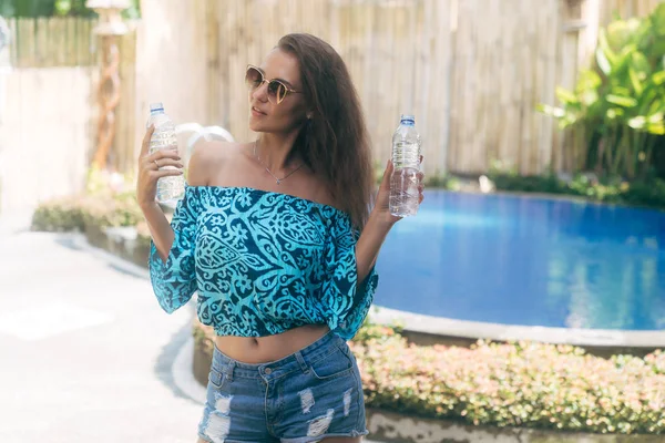 Cute girl in sunglasses and summer clothes holding two bottles of pure water on background of pool