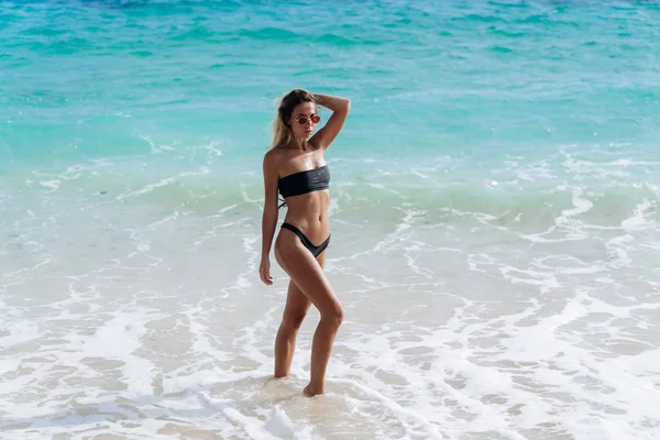 Bella ragazza in costume da bagno nero e occhiali da sole a riposo vicino all'oceano sulla spiaggia — Foto Stock