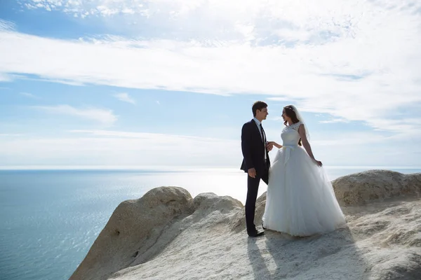 Γαμπρός στο κομψό κοστούμι και νύφη σε λευκό φόρεμα στο weddingday σε βράχο με θέα στον ωκεανό — Φωτογραφία Αρχείου