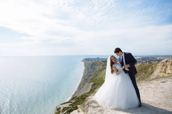 Hermosa pareja de recién casados abrazándose en el día de la boda en el acantilado con vista al mar —  Fotos de Stock