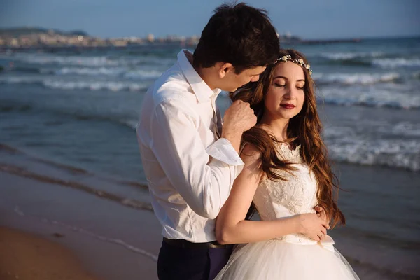 Elegant groom embraces beautiful bride in stylish wedding dress and touches her hair. Newlyweds on ocean beach during sunset time