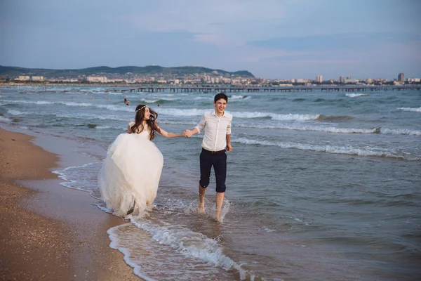 Gli sposi felici corrono lungo la riva dell'oceano. Sposi che si divertono il giorno del matrimonio sulla spiaggia tropicale — Foto Stock
