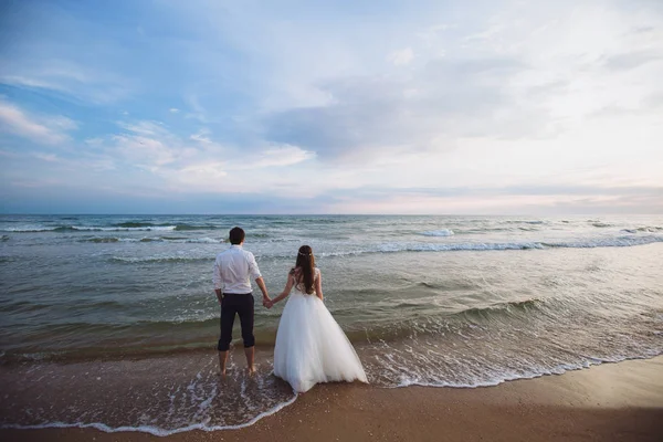 Una bella coppia di sposi novelli, gli sposi che camminano sulla spiaggia. Splendido tramonto e cielo. Abiti da sposa, un abito bianco di lusso per una ragazza. Concetto di famiglia, luna di miele . — Foto Stock