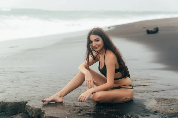 Schöne Brünette Modell im Badeanzug sitzt am schwarzen Sandstrand. — Stockfoto