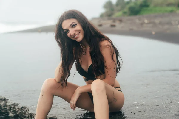 Beautiful brunette model in swimsuit sitting on black sandy beach. — Stock Photo, Image