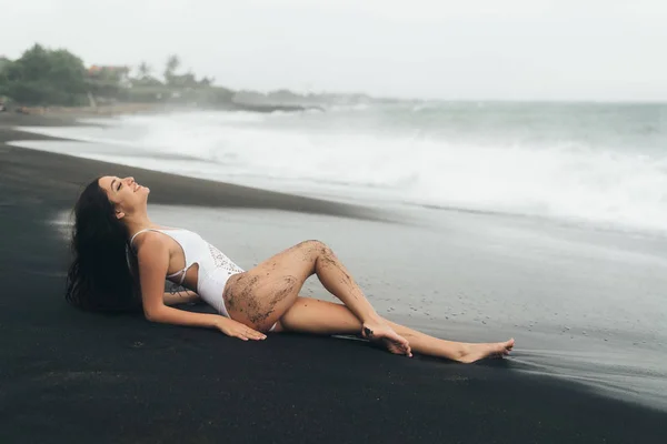 Krásná sexy dívka s dlouhými vlasy leží na pláži s černým pískem. Mladá žena se těší relaxační nedaleko oceánu — Stock fotografie