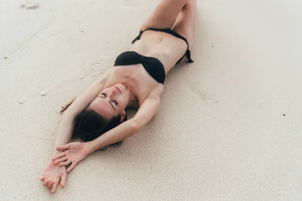 Сексуальная модель в черном купальнике, лежащая на белом песчаном пляже. Девушка загорает и отдыхает на тропическом острове — стоковое фото