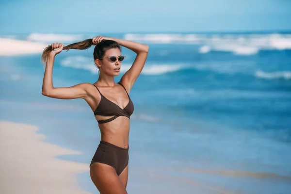 Sexy modelo bronzeado com óculos de sol em maiô marrom posando na praia de areia branca — Fotografia de Stock