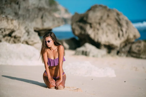 Chica bronceada sexy delgada en traje de baño posando en la playa con arena y piedras grandes —  Fotos de Stock