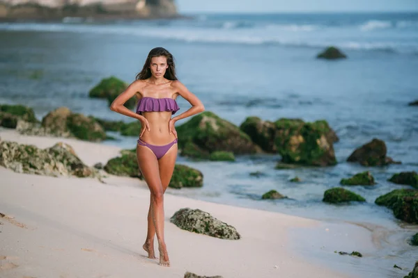Sexy žena v plavkách purpurová barva pózuje na písečné pláži s velkými kameny na pozadí — Stock fotografie