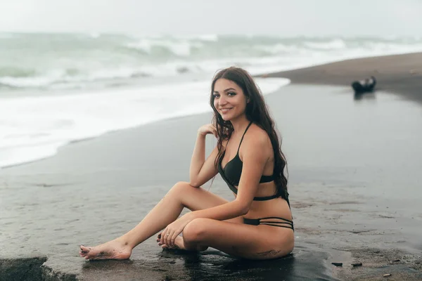 Beautiful brunette model in swimsuit sitting on black sandy beach. — Stock Photo, Image