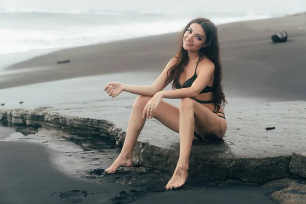 Vackra brunett modell i baddräkt som sitter på svarta sandstrand. — Stockfoto