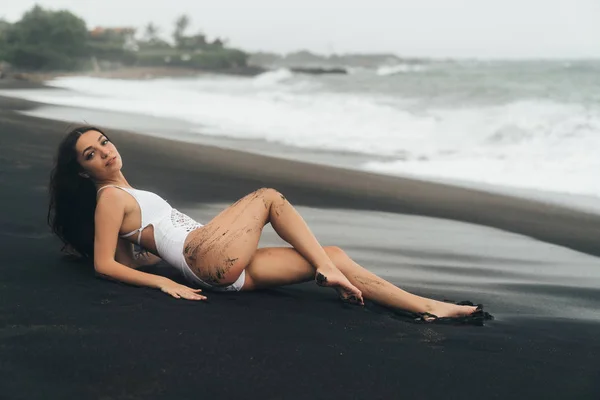 Hermosa chica sexy con el pelo largo acostado en la playa con arena negra. Mujer joven disfruta relajarse cerca del océano —  Fotos de Stock