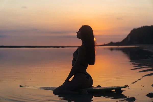 Silueta chica sexy en traje de baño acostado y posando en la tabla de surf en la playa durante la puesta del sol — Foto de Stock