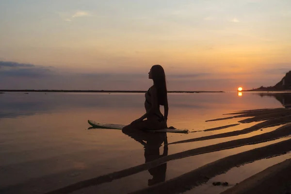 Silueta y reflejo de la niña sentada en la tabla de surf en la playa del océano en el fondo de la hermosa puesta de sol — Foto de Stock