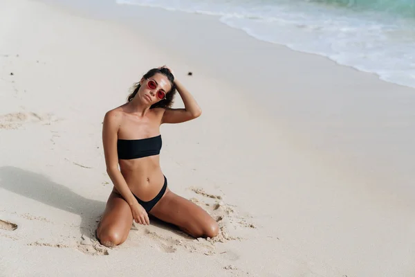 Sexy fille bronzée en maillot de bain noir et lunettes de soleil posant sur la plage de sable — Photo