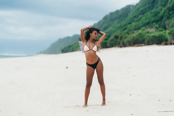 Menina esbelta sexy de pele escura em roupa de banho posando na praia com areia. Afro-americana descansando na ilha paradisíaca — Fotografia de Stock