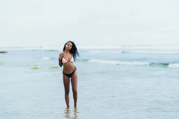 Sexy chica afroamericana en traje de baño descansando en la playa del océano. Joven mujer de piel negra con el pelo rizado se levanta en la orilla del mar — Foto de Stock