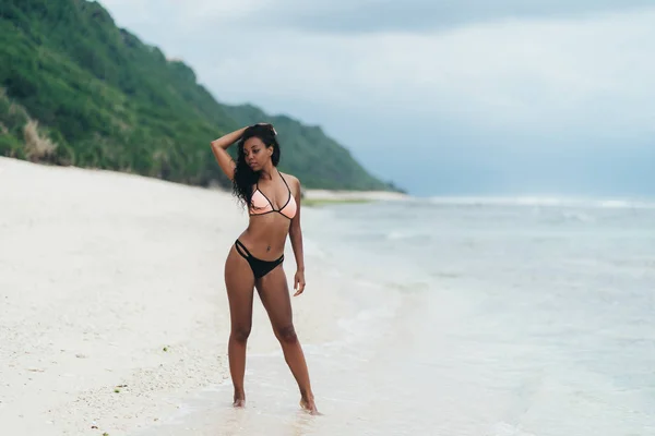 オーシャン ビーチで休んで水着でセクシーなアフリカ系アメリカ人の少女。巻き毛を持つ若い女性の黒い肌が海岸に立っています。 — ストック写真