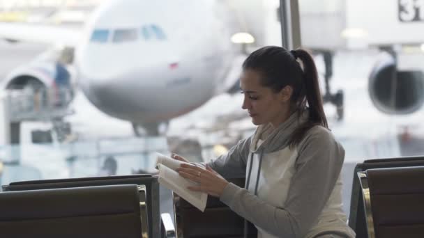 Bella ragazza si siede nel salone dell'aeroporto e legge libro sullo sfondo di aeroplano in finestra — Video Stock