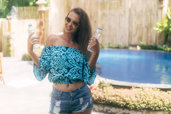 Cute girl in sunglasses and summer clothes holding two bottles of pure water on background of pool