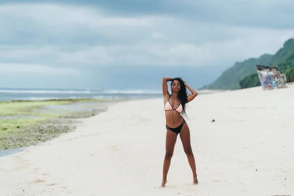 Esbelta chica sexy de piel oscura en traje de baño posando en la playa con arena. Mujer afroamericana descansando en la isla paradisíaca —  Fotos de Stock
