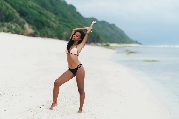 Sexy, afroamerikansk jente i badetøy som hviler på stranden. Ung, svart kvinne med krøllete hår står på land – stockfoto