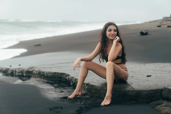 Side view of sexy girl in bikini spends time on black sand beach. Young woman with long hair posing outdoor — Stock Photo, Image