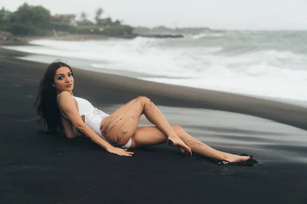 Menina encantadora com corpo bonito encontra-se na praia de areia preta. Mulher sexy descansa em férias de verão perto do oceano . — Fotografia de Stock