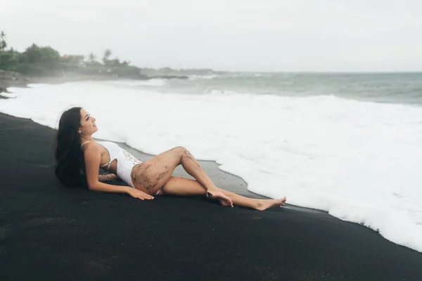 Okouzlující dívka s krásným tělem leží na pláži s černým pískem. Sexy žena se opírá o letní dovolenou u moře. — Stock fotografie