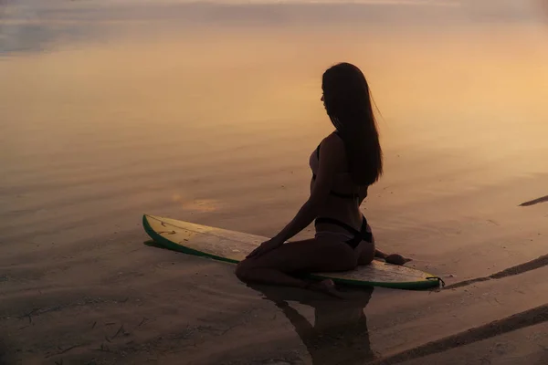 Silhouet sexy meisje in zwembroek liegen en poseren op de surfplank op strand tijdens zonsondergang — Stockfoto