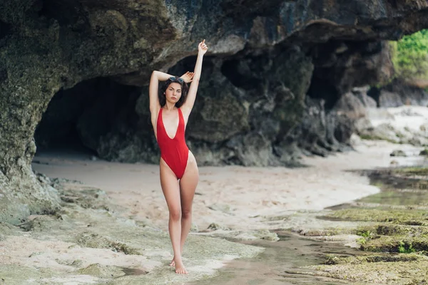 有吸引力的苗条的女孩在红色泳装摆姿势与上升的手臂附近的岩石在海滩上 — 图库照片