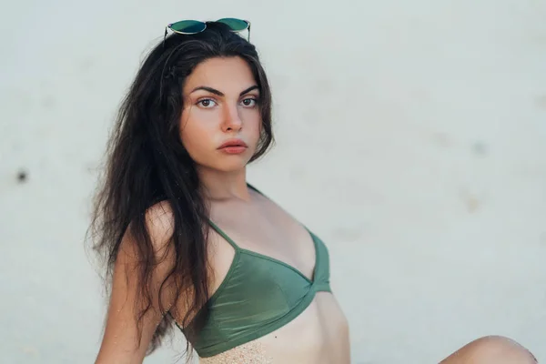 Retrato de menina sexy em roupa de banho verde e óculos de sol senta-se na praia arenosa — Fotografia de Stock